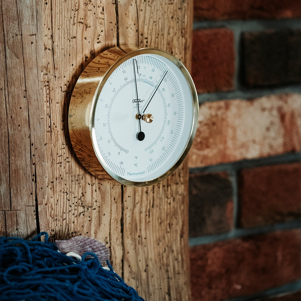 Brass wall-mounted thermometer with white dial, displayed on rustic wooden beam indoors.