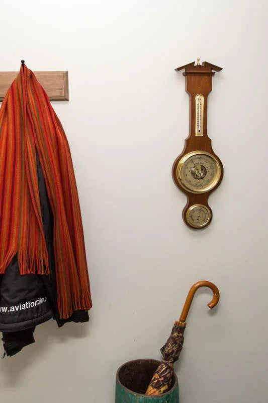 Wooden wall barometer and clock with brass dials in entryway, coat and umbrella nearby