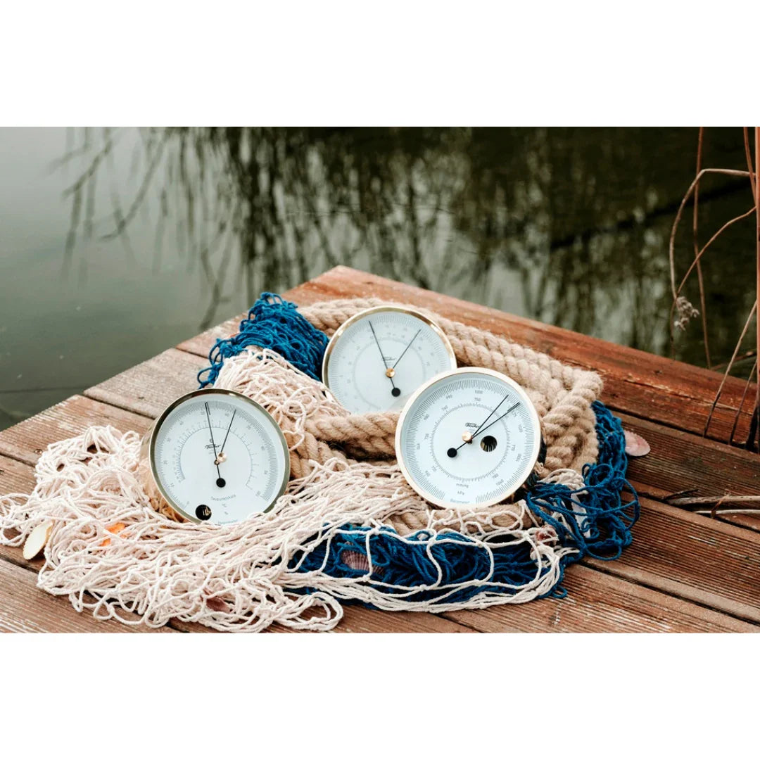 Three analog barometers and clocks on wooden dock with rope and fishing nets by water.